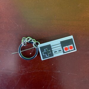 May include: A keychain featuring a miniature replica of a classic video game controller. The controller is gray and black with red buttons and a directional pad. The keychain has a silver ring and chain.