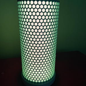 Może przedstawiać: Biała cylindryczna lampa z wzorem w kształcie plastra miodu. Lampa jest włączona i emituje zielone światło.