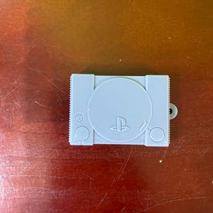 May include: A light gray PlayStation 1 console-shaped keychain. The keychain features the iconic PlayStation logo and button shapes. The design is a miniature replica of the classic gaming console, with a small loop for attaching to keys or a bag.