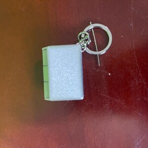 Op de afbeelding: Een kleine, rechthoekige sleutelhanger met een lichtgekleurd, getextureerd oppervlak. De sleutelhanger is bevestigd aan een zilverkleurige ring met een decoratief bloemelement. Het item ligt op een bruin oppervlak.