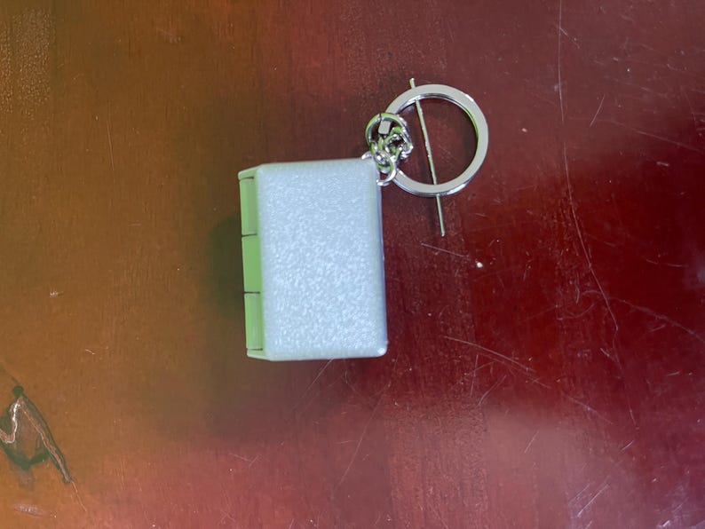 Op de afbeelding: Een kleine, rechthoekige sleutelhanger met een lichtgekleurd, getextureerd oppervlak. De sleutelhanger is bevestigd aan een zilverkleurige ring met een decoratief bloemelement. Het item ligt op een bruin oppervlak.