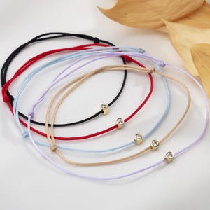 Puede incluir: Una colección de pulseras delicadas en varios colores, incluyendo negro, rojo, azul claro, lavanda y beige. Cada pulsera presenta una pequeña cuenta dorada con un acento claro y brillante. Las pulseras están dispuestas en un patrón circular.