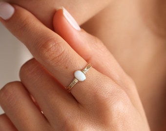 Puede incluir: Un anillo de oro con una piedra de ópalo ovalada blanca. El anillo es simple y delicado, con un diseño de alambre envuelto.