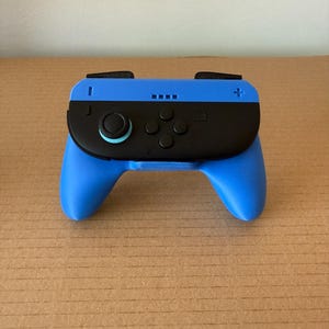 May include: A blue and black video game controller with a black joystick and buttons. The controller has a blue base and a black top section with buttons and a joystick. The controller is designed for gaming.