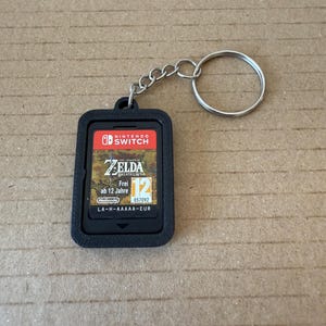Könnte beinhalten: Ein schwarzer Schlüsselanhänger mit einer Nintendo Switch-Spielkassette für "The Legend of Zelda: Breath of the Wild". Die Spielkassette befindet sich in einem schwarzen Halter und ist an einer Kette und einem Schlüsselring befestigt. Titel und Bewertung des Spiels sind sichtbar.