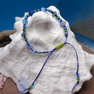 May include: A handmade macrame bracelet with blue and green beads. The bracelet has a blue cord with a green accent and a tag with the number 456. The bracelet is displayed on a white seashell.