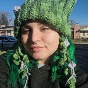 May include: A green and gray crochet hat with pointed ears and floral accents. The hat has a textured, ribbed design and is adorned with crocheted vines and lavender-colored flowers. The hat is a unique, handmade accessory.