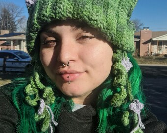 May include: A green and gray crochet hat with pointed ears and floral accents. The hat has a textured, ribbed design and is adorned with crocheted vines and lavender-colored flowers. The hat is a unique, handmade accessory.