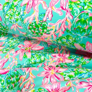 May include: A close-up of a fabric with a bright floral design. The pattern includes large pink flowers with yellow centres, green foliage, and coral-like accents on a light turquoise background. The fabric has a slight sheen and is folded.