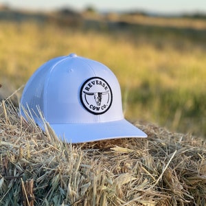 May include: White trucker hat with a black and white logo that says "Reverse Cow Co." The logo features a skull with horns.