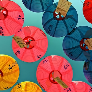May include: A collection of red, pink, blue and yellow paper lanterns hanging from the ceiling. The lanterns have white writing on them.