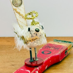 May include: A whimsical, handcrafted bird sculpture with a white body adorned with text and musical notes, perched on a red scraper. The bird features feather wings, a gold halo, and a decorative tail. The scraper has the text "Red Devil Scraper".