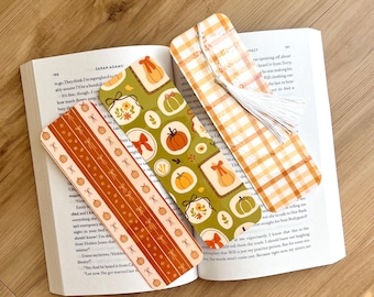 Könnte beinhalten: Drei dekorative Lesezeichen liegen auf einem offenen Buch. Die Lesezeichen zeigen herbstliche Designs, darunter Kürbisse und Karomuster. Ein Lesezeichen hat eine weiße Quaste. Die Farbpalette umfasst Orange, Grün und Weiß.
