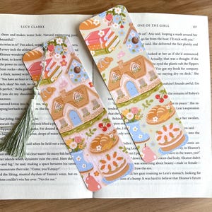 May include: Two decorative bookmarks with a cottage, flowers, bread, and cake design. The bookmarks are on a book with text. One bookmark has a green tassel.