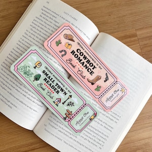 May include: Two decorative bookmarks with a ticket design are displayed on an open book. One bookmark reads "Lifetime Membership to the Cowboy Romance Book Club" and the other reads "Lifetime Membership to the Small Town Reader Book Club."