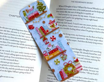 May include: A festive bookmark with a light blue background, adorned with Christmas-themed illustrations. The design includes a Christmas tree, gifts, ornaments, and the word "MERRY". The bookmark is placed on an open book.
