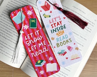 May include: Two decorative bookmarks with tassels rest on an open book. One bookmark has the text "LET IT SNOW LET ME READ" and the other says "LET'S STAY INSIDE and READ BOOKS". Both have winter-themed illustrations.