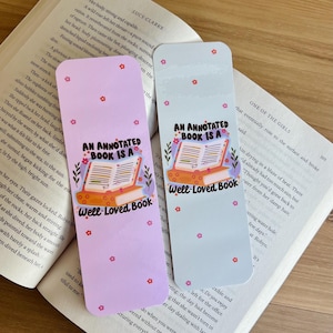 May include: Two colorful bookmarks with the text "AN ANNOTATED BOOK IS A WELL-LOVED BOOK" and floral accents. One bookmark is light purple, the other is light blue. Both are placed on an open book.