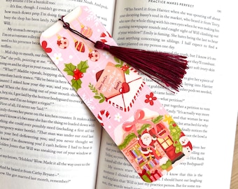May include: A pink Christmas-themed bookmark with a burgundy tassel. The bookmark features illustrations of ornaments, holly, and Santa Claus with the text "Dear Santa, All I want for Christmas is Books".