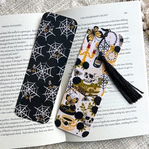 May include: Two decorative bookmarks. One black with white spiderwebs and bees. The other has a white background with skulls, butterflies, and a black tassel. The bookmarks are on top of an open book.