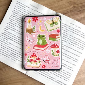 May include: Pink phone case with a whimsical design featuring a frog on a book, mushrooms, butterflies, a candle, and the text "Once Upon a Time" and "Read Dream Wander".