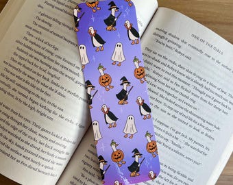 May include: A purple bookmark with a repeating pattern of Halloween-themed illustrations. The design includes penguins, ghosts, pumpkins, and witch hats. The bookmark is rectangular and is placed on top of an open book.