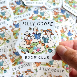 May include: A collection of stickers featuring a cartoon illustration of geese reading books. The stickers have the text "SILLY GOOSE BOOK CLUB". The geese are depicted with hats, glasses, and books in various colors. A hand holds one sticker.