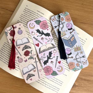 May include: Three decorative bookmarks with fantasy-themed illustrations. Each bookmark features a different design, including roses, bats, moons, books, and swords. The bookmarks have colorful tassels and are placed on an open book.