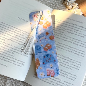 May include: A light blue bookmark with a winter theme. The bookmark features illustrations of mittens, sweaters, boots, and snowflakes. A white tassel hangs from the top. The bookmark is resting on an open book.