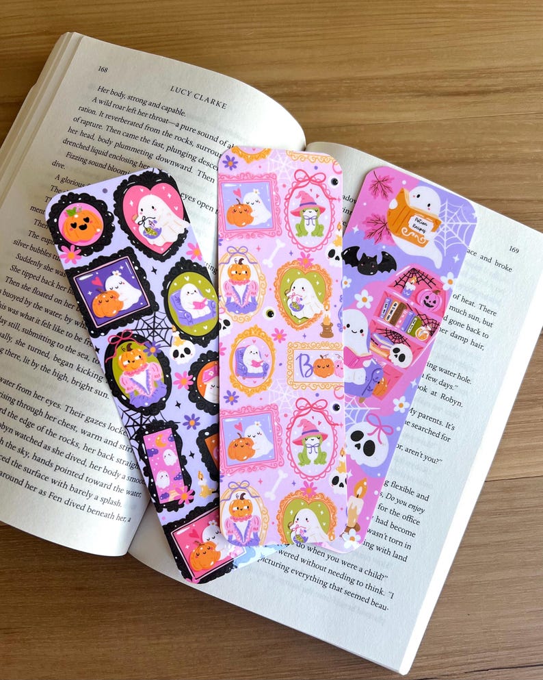 May include: Three colorful Halloween-themed bookmarks. Each bookmark features illustrations of ghosts, pumpkins, and other spooky elements within decorative frames. The bookmarks are set against an open book.