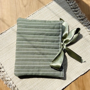 Op de afbeelding: Een groene stoffen tas met witte strepen en een satijnen lint eromheen gebonden. De tas ligt op een beige kleed met franjes.