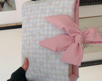 Könnte beinhalten: Ein floral gemustertes Buchcover mit hellblauem Vichy-Hintergrund und rosa Blumen. Das Cover ist mit einer großen rosa Schleife gebunden. Das Buchcover wird in einer Hand gehalten.