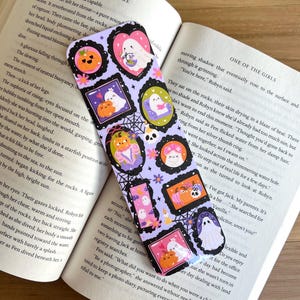 May include: A Halloween-themed bookmark featuring a purple background with various framed illustrations of ghosts, pumpkins, and spiderwebs. The bookmark is rectangular and rests on an open book.