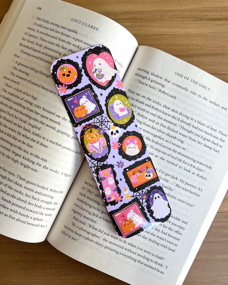 May include: A Halloween-themed bookmark featuring a purple background with various framed illustrations of ghosts, pumpkins, and spiderwebs. The bookmark is rectangular and rests on an open book.