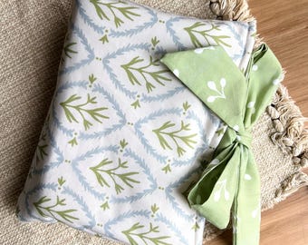 Op de afbeelding: Een wit kussen met een groen en blauw bladerpatroon, vastgebonden met een groene strik met witte bloemaccenten. Het kussen ligt op een beige geweven oppervlak met franjes, wat een zachte uitstraling creëert.