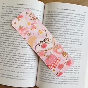 Op de afbeelding: Een roze en witte bladwijzer met herfstthema, met pompoenen, bladeren en een croissant. De bladwijzer is rechthoekig met afgeronde hoeken en ligt op een open boek.