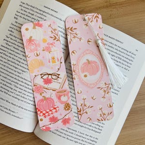 Op de afbeelding: Twee roze bladwijzers met herfstthema-illustraties. E&eacute;n heeft een pompoen in een lijst, de andere heeft pompoenen, bladeren en een kopje koffie. Beide hebben een witte kwast en liggen op een open boek.