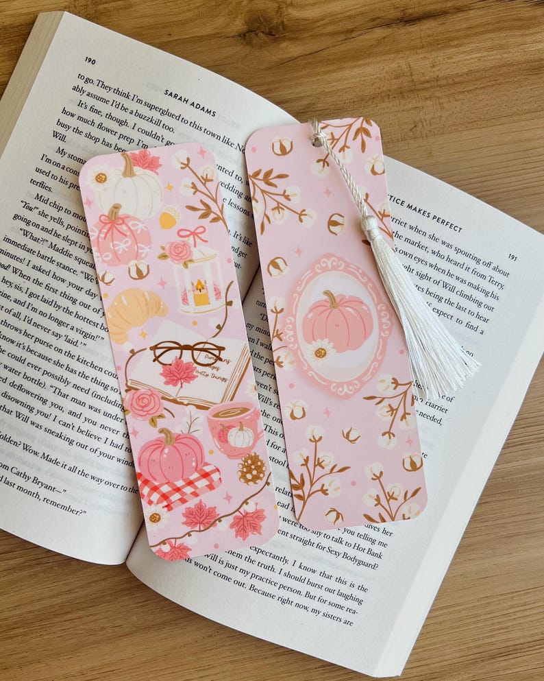 Op de afbeelding: Twee roze bladwijzers met herfstthema-illustraties. E&eacute;n heeft een pompoen in een lijst, de andere heeft pompoenen, bladeren en een kopje koffie. Beide hebben een witte kwast en liggen op een open boek.