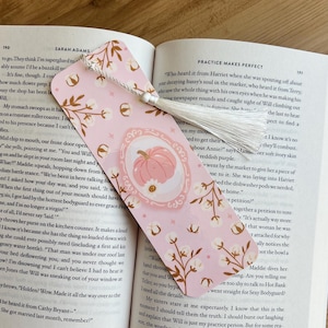 Op de afbeelding: Roze bladwijzer met een pompoenillustratie in een ovaal kader, omringd door katoentakontwerpen. De bladwijzer heeft een witte kwast en ligt in een open boek.