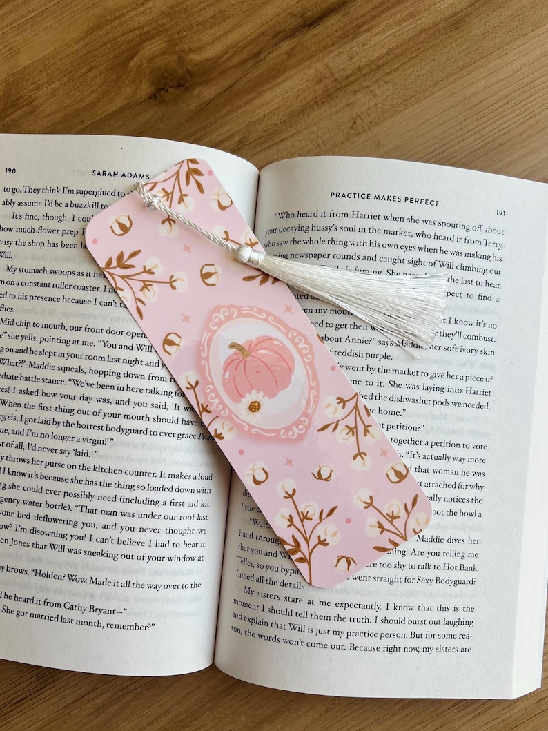 Op de afbeelding: Roze bladwijzer met een pompoenillustratie in een ovaal kader, omringd door katoentakontwerpen. De bladwijzer heeft een witte kwast en ligt in een open boek.