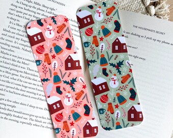 May include: Two decorative bookmarks with a winter holiday theme. One is pink, the other is green. Both feature a repeating pattern of houses, snowmen, mittens, hats, and holly. The bookmarks are on top of an open book.