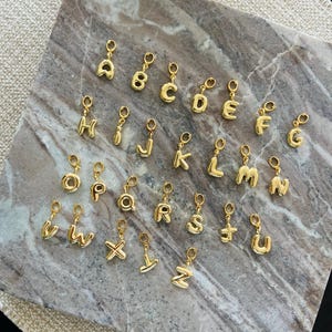 May include: Gold alphabet letter charms with a jump ring for attaching to a necklace or bracelet. The letters are in a bubble font style.