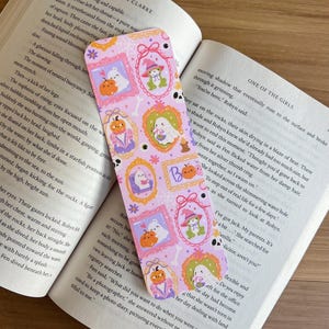 May include: A decorative bookmark with a light purple background featuring framed illustrations of ghosts, pumpkins, and a frog wearing a witch hat. The bookmark is placed in an open book.