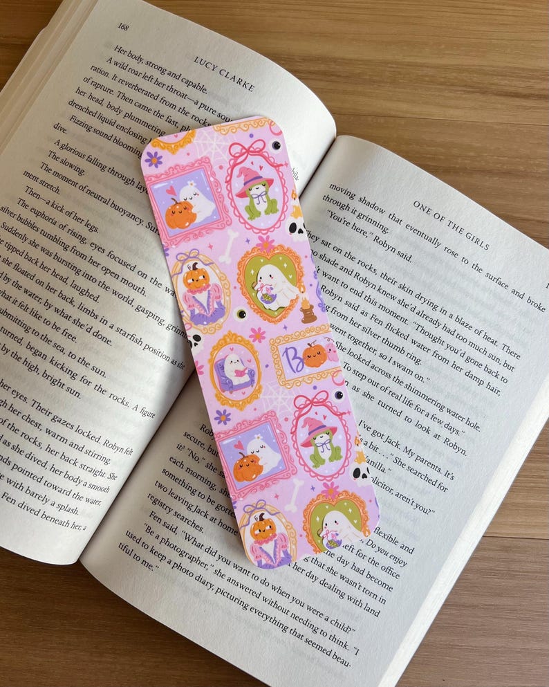 May include: A decorative bookmark with a light purple background featuring framed illustrations of ghosts, pumpkins, and a frog wearing a witch hat. The bookmark is placed in an open book.