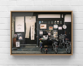 May include: A storefront with a sign that reads "Gallery Kaiguraku", a cafe, and a sign that reads "Cafe Hinterbeck". There are bicycles parked in front of the store.