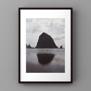 May include: A framed wall art print featuring a dark, monolithic sea stack rising from a wet, reflective beach under a cloudy sky. The artwork is encased in a dark brown rectangular frame with a wide white mat border surrounding the photograph.