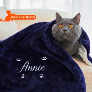 Può includere: Una coperta in pile blu navy con fodera in sherpa bianca. La coperta ha impronte di zampe bianche e il nome "Annie" ricamato. Un gatto grigio è rannicchiato sulla coperta.