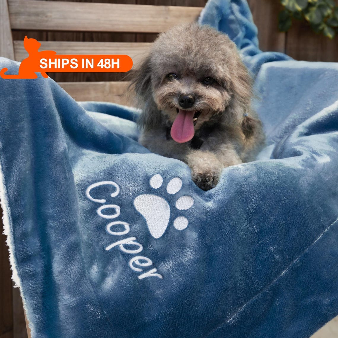 A small brown dog lying on a blue blanket embroidered with the name Cooper and a paw print.