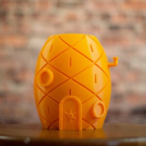 May include: A yellow pineapple-shaped ceramic planter with a small door and two round windows. The planter is sitting on a wooden surface.