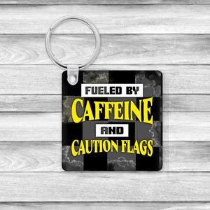 May include: Square keychain with a silver ring. The keychain features a black and white chequered pattern with a cloudy design. The text on the keychain reads "Fueled by Caffeine and Caution Flags" in yellow and white.